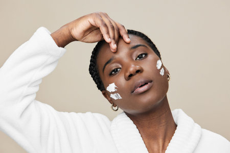 Young Black woman applying skincare product with a serene expression, dressed in a white bathrobe against a soft beige background, highlighting beauty and self careの写真素材