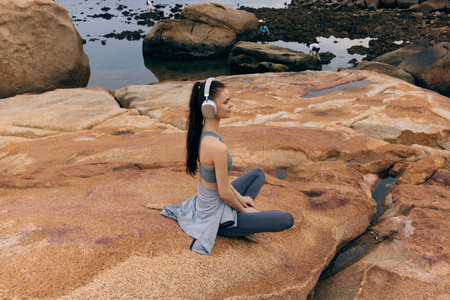 Young woman practicing meditation on rocky shore, wearing sportswear and headphones, showcasing tranquility amidst natureの写真素材