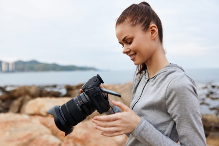 Young woman reviewing photos on her camera by the ocean, wearing a casual gray hoodie, showcasing joy and creativity in a serene coastal settingの写真素材