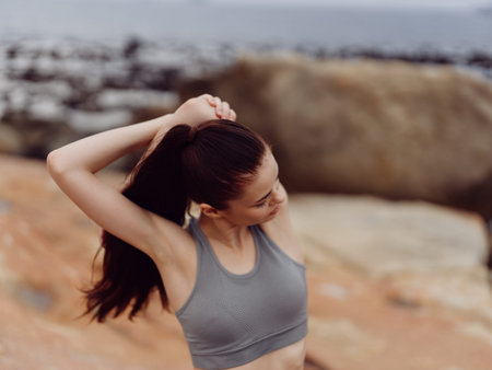 Young woman stretching outdoors on a rocky beach, wearing a gray sports bra and enjoying a moment of...