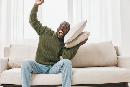 Joyful African man celebrating at home, holding pillows in a cozy living room, wearing a green sweater and denim jeans, conveying happiness and excitementの写真素材