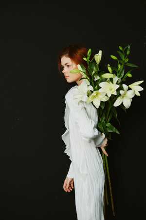 Elegant woman with red hair in a white dress holding white lilies against a dark background,...