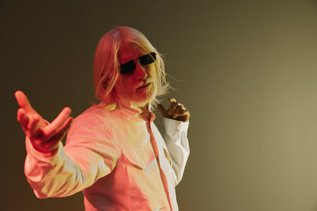 Stylish older man with white hair and sunglasses in a modern white shirt striking a pose against a gradient colored background, showcasing confidence and charismaの写真素材