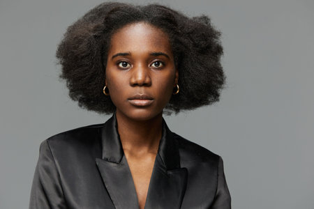 Portrait of a confident Black woman with natural hair, wearing a stylish black satin blazer, showcasing strength and elegance against a minimalist gray backgroundの写真素材