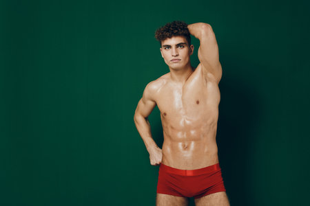 Confident young man posing in red shorts, showcasing toned physique against a solid green background. Fitness and health concept.の写真素材