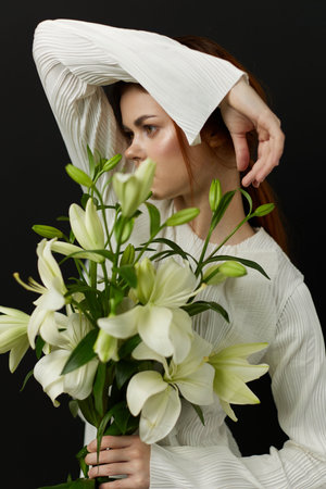 Young woman in a white outfit holding white lilies against a dark background, expressing serenity and elegance in a floral compositionの写真素材