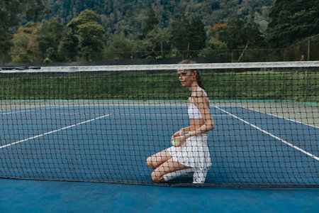Young female tennis player in a white outfit kneeling on a blue tennis court, looking determined and focused, surrounded by nature and greeneryの写真素材