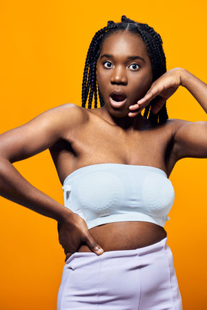 Shocked young woman with braided hair in a strapless top and pastel pants, posing against a vibrant yellow background, expressing surprise and excitementの写真素材