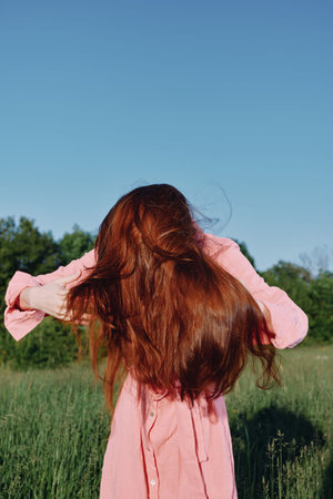 Young woman with flowing red hair in a pink dress enjoying nature against a green backdrop, representing freedom and natural beautyの写真素材