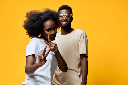Young Afro American couple posing playfully with peace signs against a vibrant yellow background,...