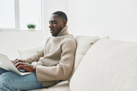 A young Black man in a cozy beige sweater working on a laptop while sitting on a light sofa, showcasing focus and productivity at homeの写真素材