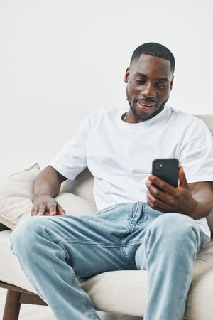 Happy young African man in casual outfit sitting on a sofa and using smartphone, enjoying social...