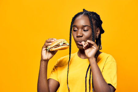 Young girl enjoying a delicious burger, wearing a bright yellow shirt, set against a vibrant yellow background, showcasing joy and a love for foodの写真素材