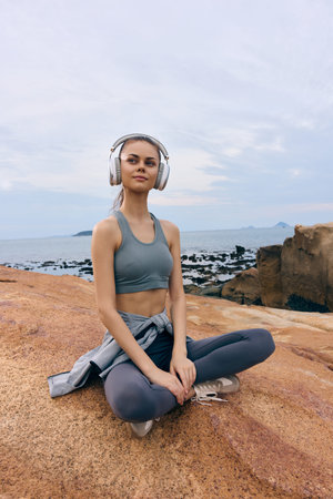 Young woman practicing mindfulness and meditation on rocky beach, wearing sportswear and headphones,...