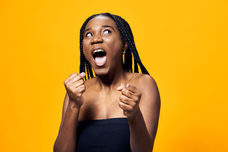 Excited young woman expressing joy with raised fists on a vibrant yellow background, showcasing...