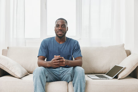 Young African man smiling confidently while sitting on a couch with a laptop, portraying a relaxed and inviting atmosphere in a bright roomの写真素材
