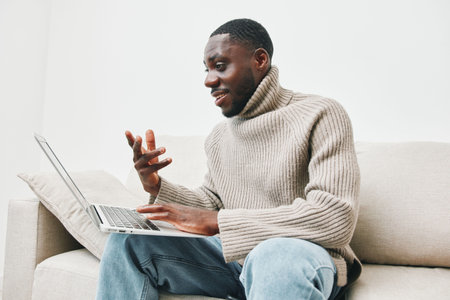 Happy African man in cozy sweater using laptop for online communication at home, enjoying comfortable lifestyle and technology connectionの写真素材