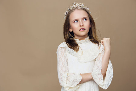 A young girl with a crown, wearing a white lace dress, gazes thoughtfully against a beige background, exuding innocence and eleganceの写真素材