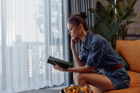 Cozy woman enjoying a book and a meal on an orange couch in a comfortable living room settingの写真素材