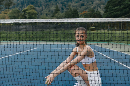 Young athletic woman in sportswear sitting by the tennis net, showcasing a confident and determined expression on a sunny day on the courtの写真素材