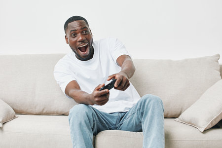 Excited young African man playing video games on a cozy couch, showcasing joy and engagement in a...