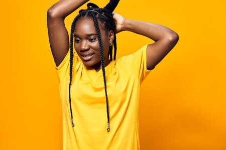 Young Black woman with braided hair smiling while adjusting her hair against a vibrant yellow...