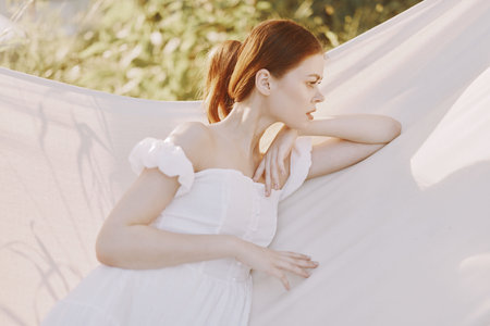 A serene young woman in a white dress relaxes in a hammock, surrounded by soft greenery and warm sunlight, embodying tranquility and summer vibesの写真素材