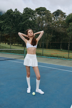 Athletic young woman in a white tennis outfit poses confidently on a tennis court, showcasing sportswear style and a vibrant outdoor atmosphereの写真素材
