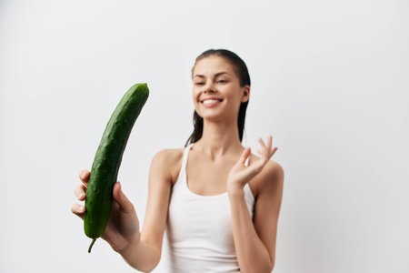 Smiling woman holding a cucumber in a bright, minimalist setting A healthy concept emphasizing fresh produce and wellness The background is a smooth, light colorの写真素材