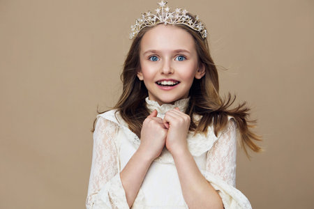 A joyful young girl with a crown, dressed in a vintage white dress, smiling brightly against a neutral beige background, representing innocence and childhood wonderの写真素材