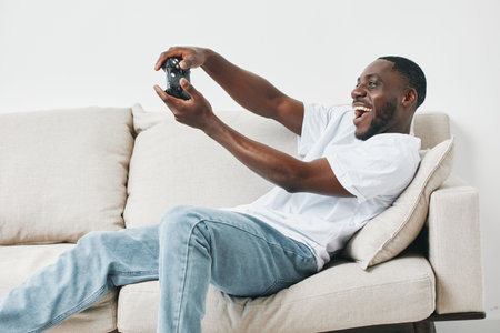 Joyful young African American man playing video games on the couch, wearing a casual white t shirt and jeans, exhibiting excitement and happiness against a neutral backgroundの写真素材