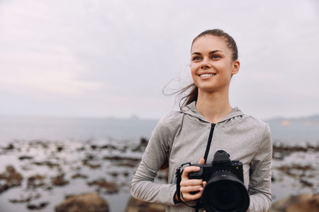 A smiling young woman with long hair in a gray sports hoodie holds a camera, enjoying her time by...