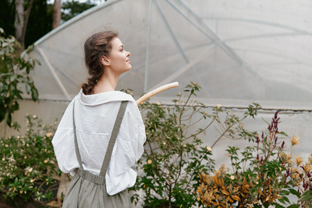 Young woman in a greenhouse wearing casual outfit, smiling while holding gardening tool surrounded by lush plants, represents sustainable living and gardening passionの写真素材