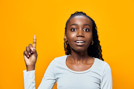 Young Black girl with curly hair raises her finger in excitement against a vibrant yellow background, showcasing a moment of inspiration and joyの写真素材