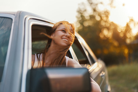 Young woman enjoying a summer road trip, smiling joyfully with wind in her hair while looking out of the car window in a lush green landscape.の写真素材
