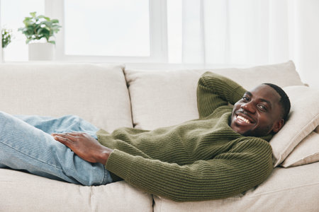 Smiling young African man relaxing on a couch in a cozy living room, wearing a green sweater, capturing a warm and inviting atmosphereの写真素材