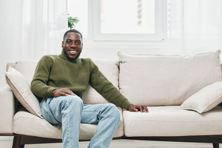 Smiling African American man in a green sweater relaxing on a beige sofa, enjoying a bright and cozy indoor setting, adding warmth and comfort to home decorの写真素材