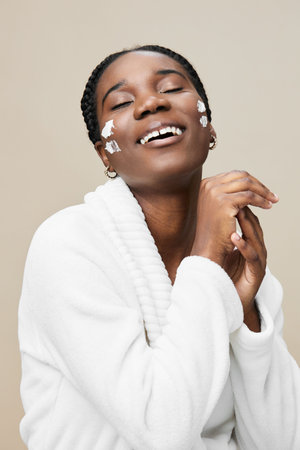 Joyful black woman with skincare cream on her face, wearing a cozy white robe, smiling in a soft toned background, emphasizing beauty and self careの写真素材