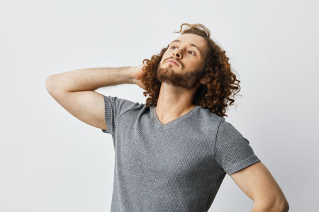 Confident young man with curly hair in casual gray t shirt showcasing self assurance and style against a neutral backgroundの写真素材