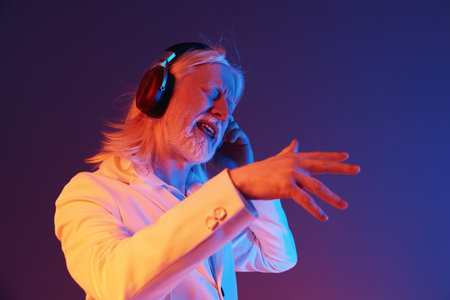 Elderly man enjoying music, wearing headphones, in a vibrant colorful background with gradient hues of blue and purple, conveying emotions of joy and celebration through musicの写真素材