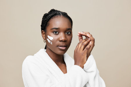 Young African woman applying facial cream, showcasing natural beauty and skincare routine, calm expression, minimalistic background, soft tones for beauty conceptsの写真素材