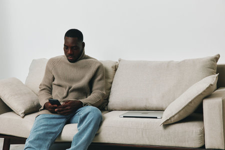 Young Black man sitting on a beige couch, checking his phone while relaxed, portraying a sense of calm and modern lifestyle in a minimalist environmentの写真素材