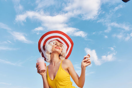 Happy woman enjoying a drink under a sunny sky with fluffy clouds and a large striped hatの写真素材
