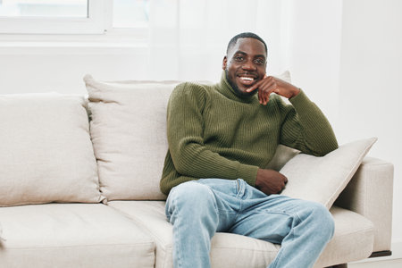 A smiling African man in a green turtleneck sweater seated on a light couch in a bright, modern living room, exuding a cozy and relaxed atmosphereの写真素材