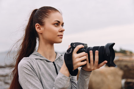 Young female photographer holding a camera near the beach, dressed in a gray hoodie, showcasing focus and creativity with a serene ocean backdropの写真素材