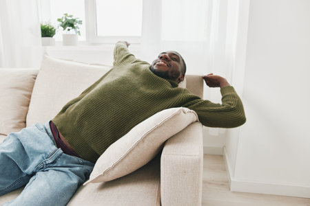 Relaxed young African man enjoying leisure time at home in cozy sweater and jeans, expressing comfort and tranquility in a bright, minimalistic living roomの写真素材