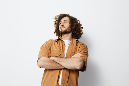 Confident young man with curly hair in a casual brown shirt, standing against a white background, showcasing a thoughtful expression and relaxed poseの写真素材