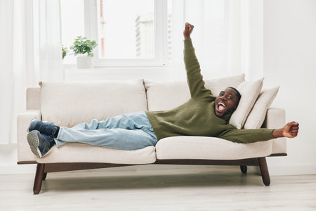 A joyful Black man relaxing on a cozy sofa, celebrating life with wide smiles and open arms, in a bright and minimalist living roomの写真素材