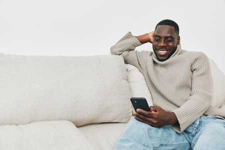 Relaxed Black man smiling while using a smartphone on a cozy couch, showcasing casual style and comfort in a modern living spaceの写真素材
