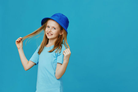Cheerful girl with long hair in a blue hat, smiling and playing with her hair against a vibrant blue background, perfect for children s fashion themesの写真素材